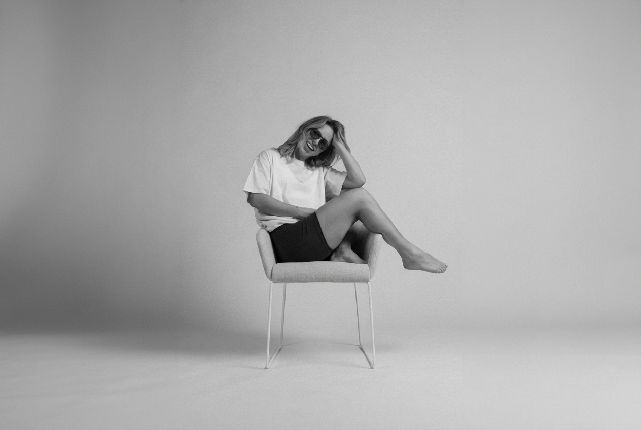 A woman in an oversized white t-shirt sitting on a light grey chair, smiling behind sunglasses